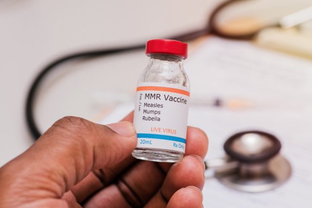 measles mumps and rubella vaccination concept with african american doctor hand holding vaccine vial and stethoscope and syringe and checklist in background